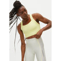 A person with long braids wears a yellow Kay Sports Bra by Rohnisch, crafted from moisture-wicking fabric, along with grey athletic pants, adjusting the waistband of the pants. The background is plain white.