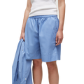 A person is wearing a white sleeveless top and Aiayu's blue GOTS-certified Shorts Long with an elastic waistband, holding a blue shirt in their left hand.