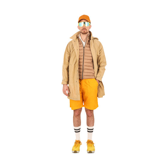 A man stands sporting an orange cap, reflective sunglasses, a beige jacket, and Universal Works' Pleated Track Short in orange. His vibrant look is completed with striped socks and yellow sneakers.