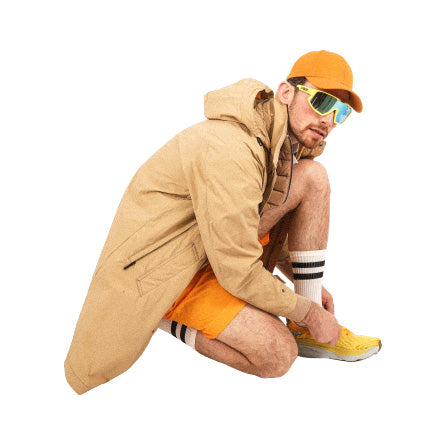 A person in a tan coat, orange cap, and Universal Works Pleated Track Short in Orange crouches to adjust their yellow shoes, wearing reflective sunglasses.