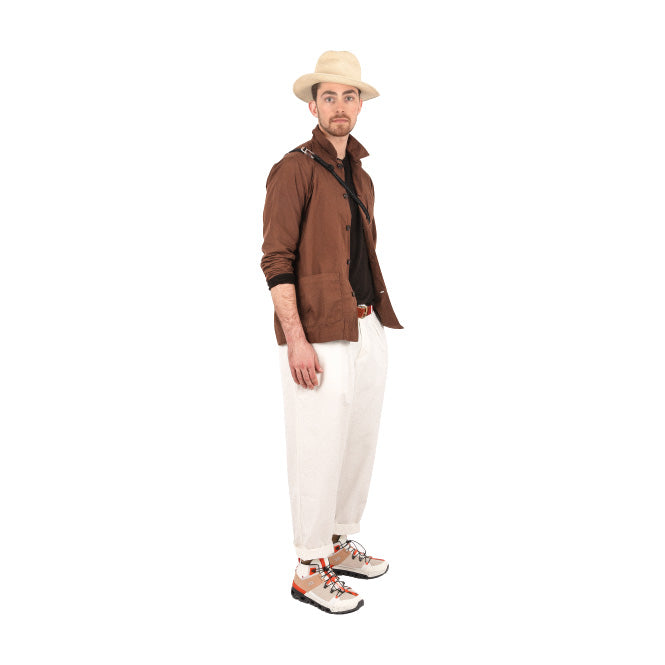 A man in a brown jacket and black shirt sports Universal Works' Oxford Pant in Ecru, featuring a single pleat and tapered leg. He wears white sneakers with orange accents and completes his look with a beige hat against a crisp white background.